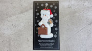 Ornament of white snowman in red hat holding a briefcase and paper bills, shown on backing card and Christmas tree.