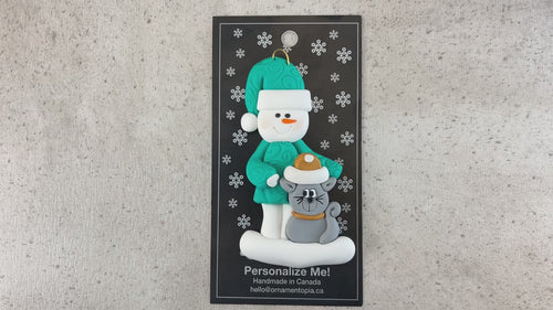 Ornament of snowman in a green shirt and hat with grey cat in Christmas hat, shown on backing card and Christmas tree.