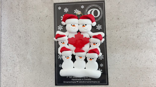 Family ornament with 7 white snowmen in red hats, gathered around a red maple leaf, shown on backing card and Christmas tree.