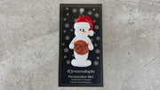 Ornament of white snowman in red hat holding a basketball, shown on backing card and Christmas tree.