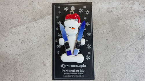 Ornament of white snowman in red hat holding a pair of vibrant skis, shown on backing card and Christmas tree.