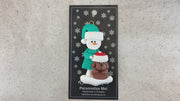 Ornament of snowman in green shirt and hat with brown dog in Christmas hat, shown on backing card and Christmas tree.