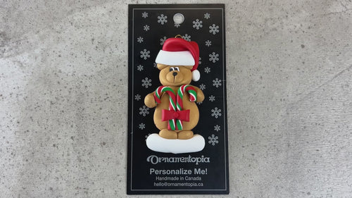 Tan bear in red festive hat holding two candy canes ornament, shown on backing card and Christmas tree.