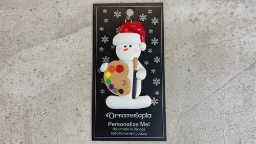 Ornament of white snowman in red hat holding a paint palette and paintbrush, shown on backing card and Christmas tree.