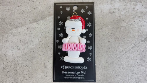 Ornament of white snowman in red hat wearing a pink tutu, shown on backing card and Christmas tree.