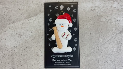 Ornament of white snowman in red hat holding a baseball bat and a baseball, shown on backing card and Christmas tree.