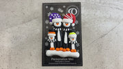 Family ornament with 4 black and white penguins in colourful festive hats, shown on backing card and Christmas tree.