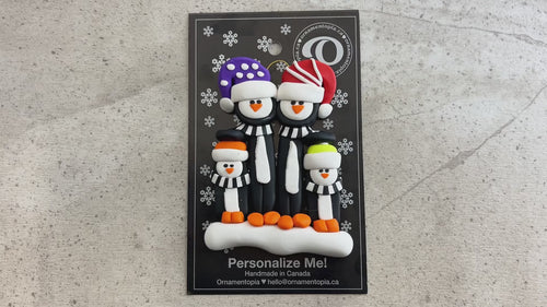 Family ornament with 4 black and white penguins in colourful festive hats, shown on backing card and Christmas tree.