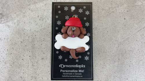 Ornament of brown dog in red hat hanging on a white bone, shown on backing card and Christmas tree.