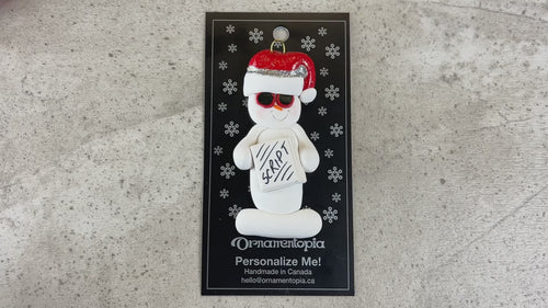 Ornament of white snowman in red hat and sunglasses holding a movie script, shown on backing card and Christmas tree.