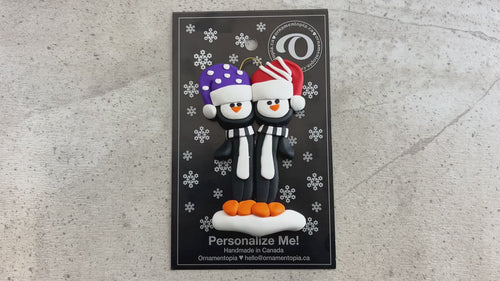 Family ornament with 2 black and white penguins in colourful festive hats, shown on backing card and Christmas tree.