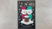 Ornament of snowman couple in red and green shirts and hats with brown dog in Christmas hat, shown on backing card and Christmas tree.