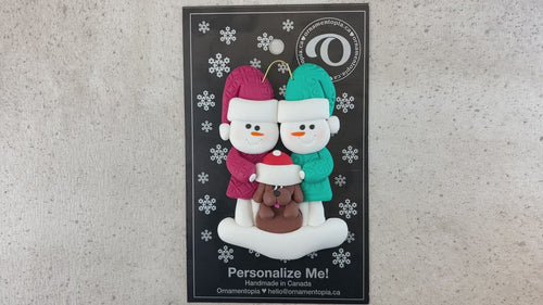 Ornament of snowman couple in red and green shirts and hats with brown dog in Christmas hat, shown on backing card and Christmas tree.