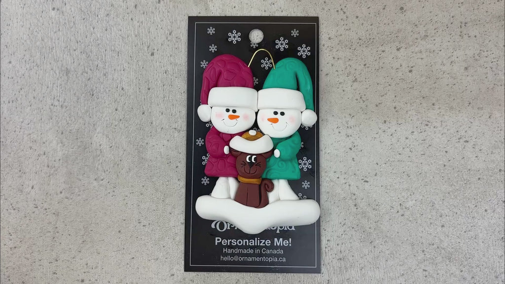 Ornament of snowman couple in red and green shirts and hats with a brown cat in Christmas hat, shown on backing card and Christmas tree.