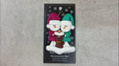Ornament of snowman couple in red and green shirts and hats with a brown cat in Christmas hat, shown on backing card and Christmas tree.