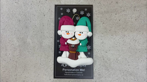Ornament of snowman couple in red and green shirts and hats with a brown cat in Christmas hat, shown on backing card and Christmas tree.