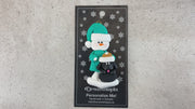 Ornament of snowman in green shirt and hat with black dog in Christmas hat, shown on backing card and Christmas tree.