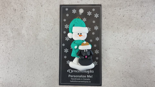 Ornament of snowman in green shirt and hat with black dog in Christmas hat, shown on backing card and Christmas tree.