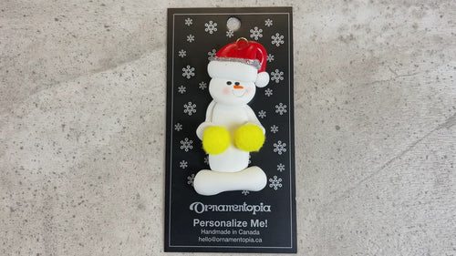 Ornament of white snowman in red hat holding yellow cheerleader pom-poms, shown on backing card and Christmas tree.