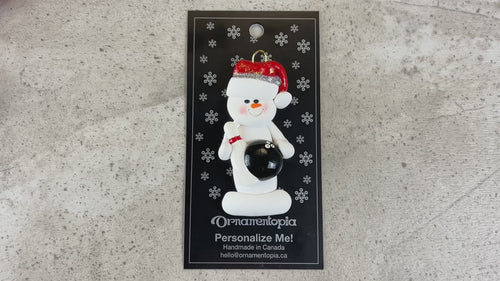 Ornament of white snowman in red hat holding a black bowling ball and pin, shown on backing card and Christmas tree.