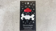 Ornament of black dog in red hat hanging on a white bone, shown on backing card and Christmas tree.