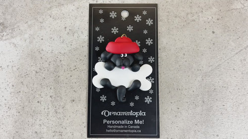 Ornament of black dog in red hat hanging on a white bone, shown on backing card and Christmas tree.