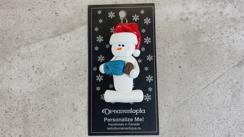 Ornament of white snowman in red hat holding colourful mug and coffee bean, shown on backing card and Christmas tree.