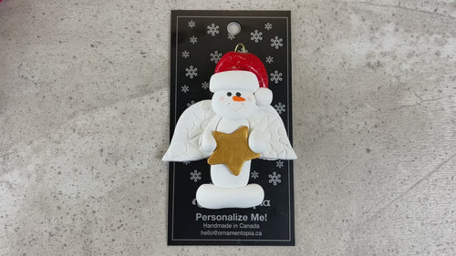 Ornament of white snowman in red hat with angel wings and a golden star, shown on backing card and Christmas tree.