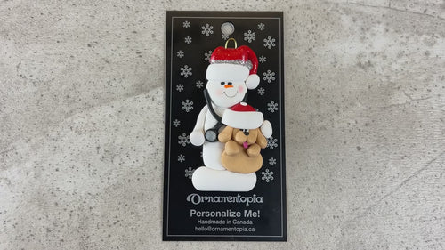 Ornament of white snowman in red hat holding a tan dog and stethoscope, shown on backing card and Christmas tree.