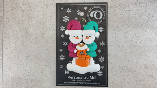 Ornament of snowman couple in red and green shirts and hats with a orange cat in Christmas hat, shown on backing card and Christmas tree.