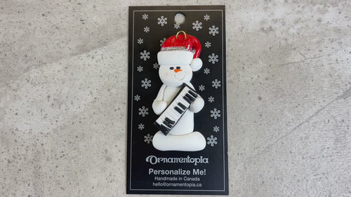 Ornament of white snowman in red hat holding a piano keyboard, shown on backing card and Christmas tree.