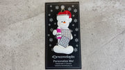 Ornament of white snowman in red hat holding a bingo card and dabber, shown on backing card and Christmas tree.