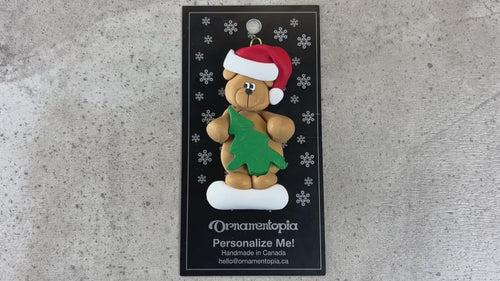 Tan bear in red festive hat holding green tree ornament, shown on backing card and Christmas tree.