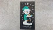 Ornament of snowman in a green shirt and hat with black cat in Christmas hat, shown on backing card and Christmas tree.