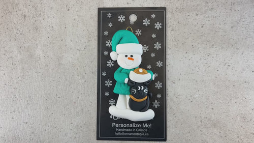 Ornament of snowman in a green shirt and hat with black cat in Christmas hat, shown on backing card and Christmas tree.
