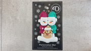 Ornament of snowman couple in red and green shirts and hats with tan dog in Christmas hat, shown on backing card and Christmas tree.