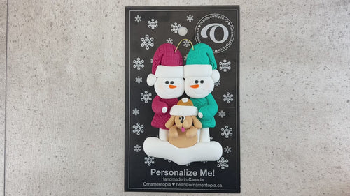 Ornament of snowman couple in red and green shirts and hats with tan dog in Christmas hat, shown on backing card and Christmas tree.