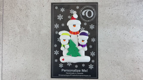 Family ornament with 1 large and 2 small white snowmen in bright scarves, with green Christmas tree, shown on backing card and Christmas tree.