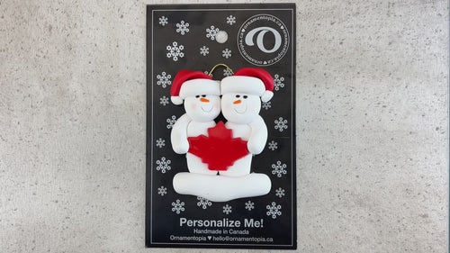 Family ornament with 2 white snowmen in red hats, gathered around a red maple leaf, shown on backing card and Christmas tree.