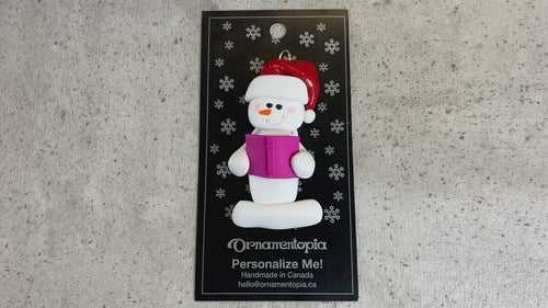 Ornament of white snowman in red hat holding a pink book, shown on backing card and Christmas tree.