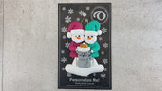 Ornament of snowman couple in red and green shirts and hats with grey cat in Christmas hat, shown on backing card and Christmas tree.
