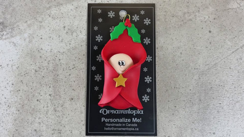 Ornament with an infant head poking out of a red baby blanket, shown on backing card and Christmas tree.