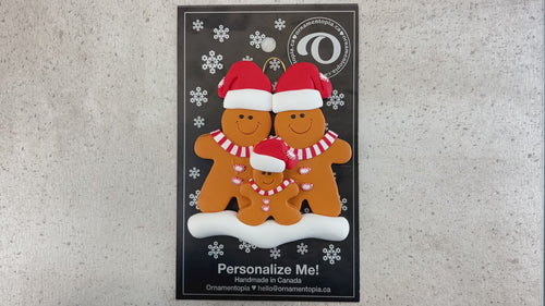 Family ornament with 3 brown gingerbread figures in red Christmas hats, shown on backing card and Christmas tree.