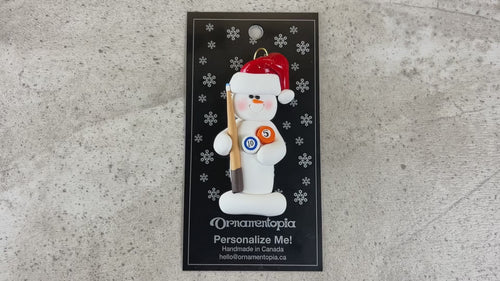Ornament of white snowman in red hat holding a pool cue and pool balls, shown on backing card and Christmas tree.