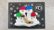 Family ornament with 4 white snowmen in bright hats, holding a hockey stick, shown on backing card and Christmas tree.
