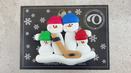 Family ornament with 4 white snowmen in bright hats, holding a hockey stick, shown on backing card and Christmas tree.