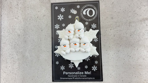 Family ornament with 5 white snowmen stacked on top of a glitter white snowflake, shown on backing card and Christmas tree.