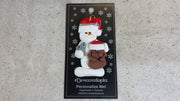 Ornament of white snowman in red hat holding a brown dog and a pair of scissors, shown on backing card and Christmas tree.