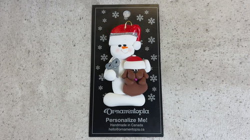 Ornament of white snowman in red hat holding a brown dog and a pair of scissors, shown on backing card and Christmas tree.
