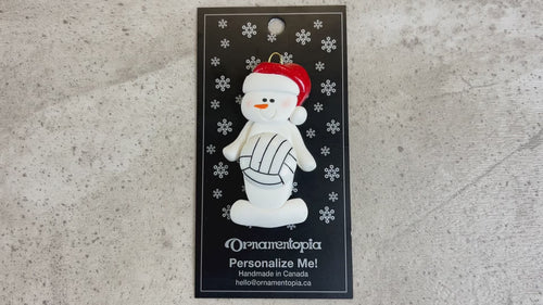 Ornament of white snowman in red hat holding a volleyball, shown on backing card and Christmas tree.